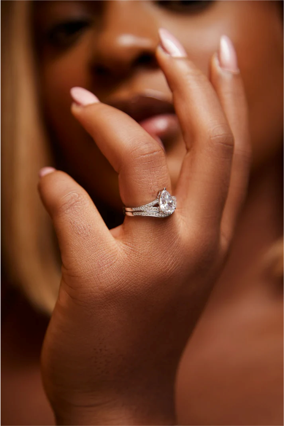 A close-up of a person's hand showcasing a gold ring with a pear cut Moissanite center stone and diamond side stones.
