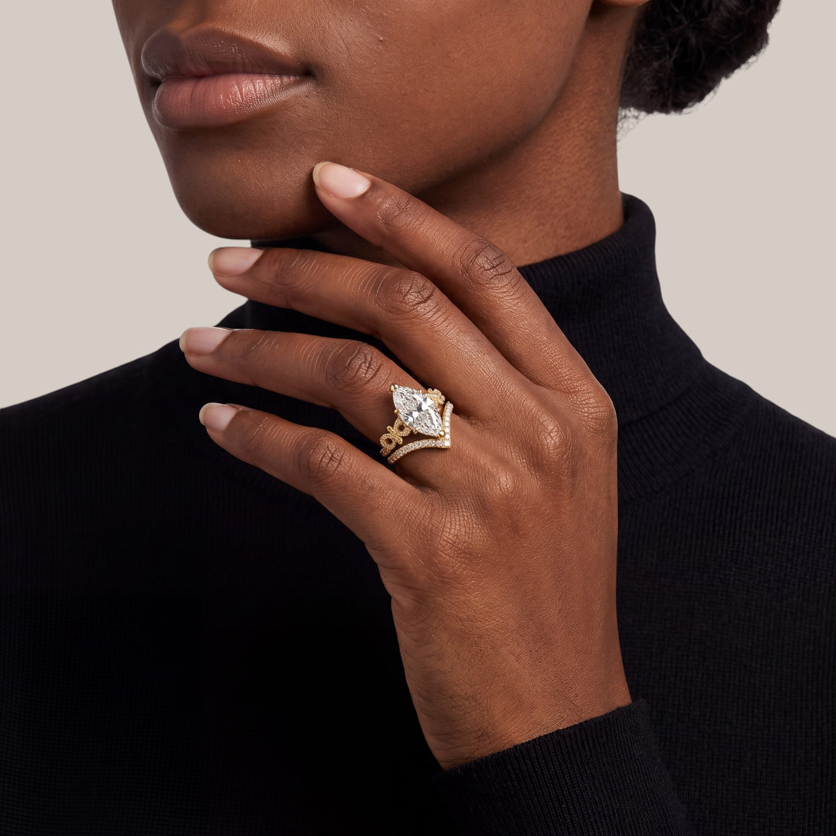 A close-up of a model's hand wearing a gold ring with a marquise-cut clear gemstone and smaller accent stones, paired with a black turtleneck sweater against a neutral beige background.