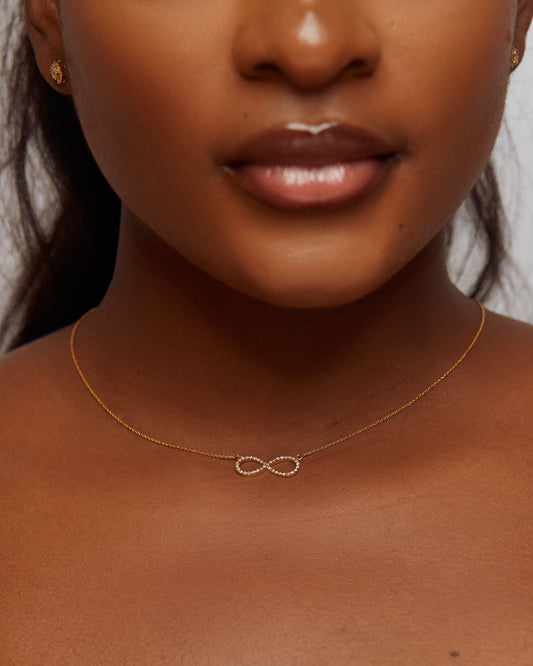 Close-up of a person wearing a gold necklace with an infinity symbol on a neutral background