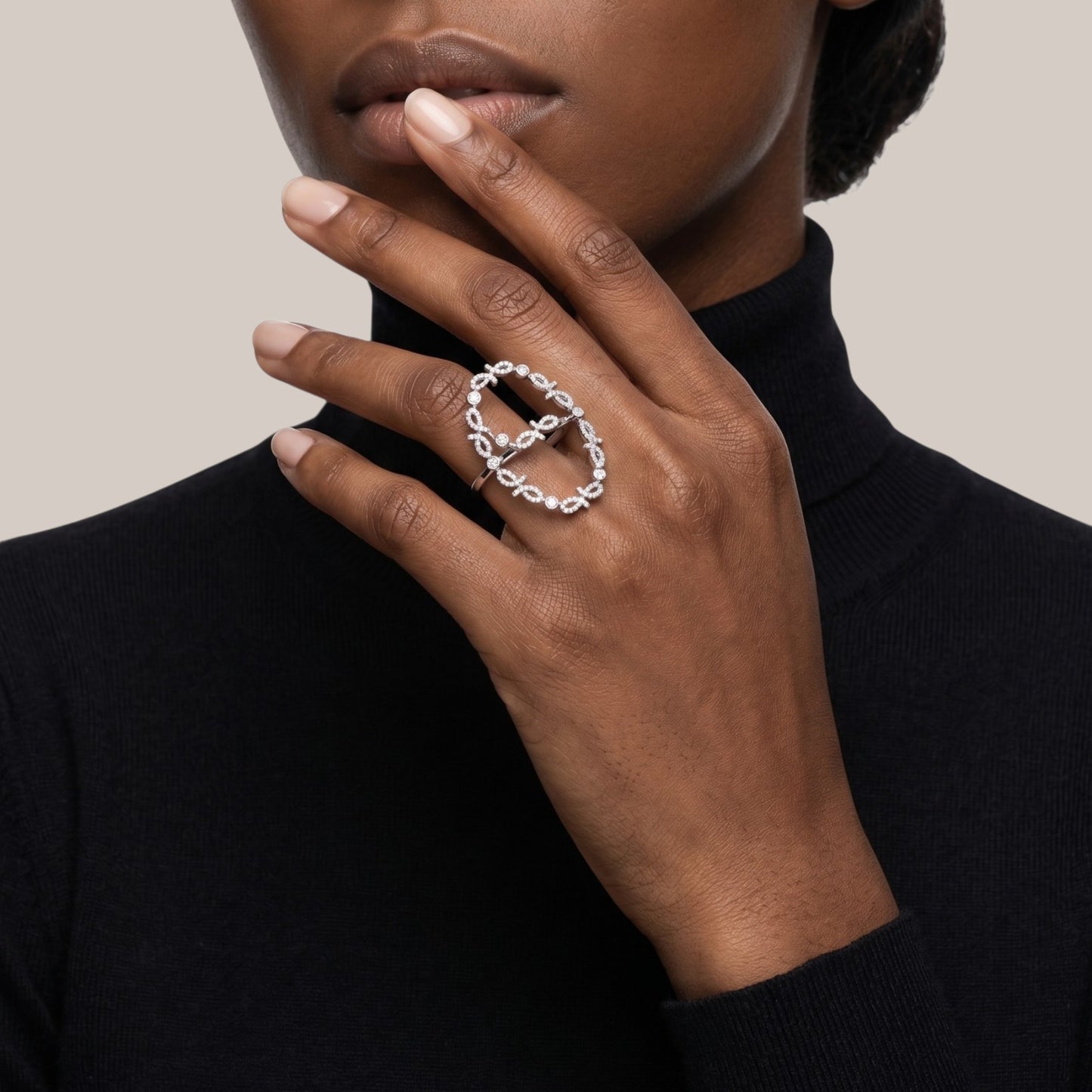 A close-up of a model's hand wearing a silver infinity ring with a central cross, styled with a black turtleneck against a neutral beige background.