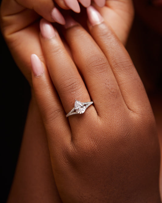 A close-up image of a woman's hand holding a pear cut cubic zirconia engagement ring with a split shank design, set in 10 karat gold.