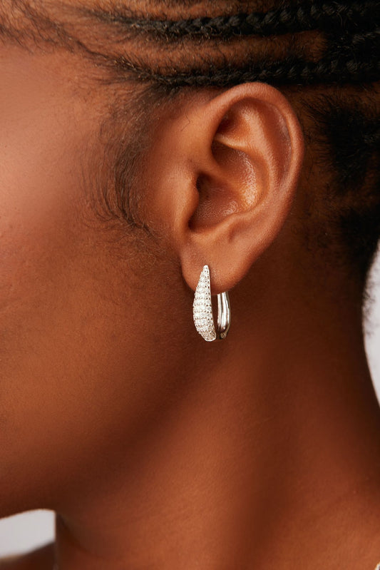 A close-up image of a woman's ear wearing a silver hoop earring with a clear, sparkling gemstone.